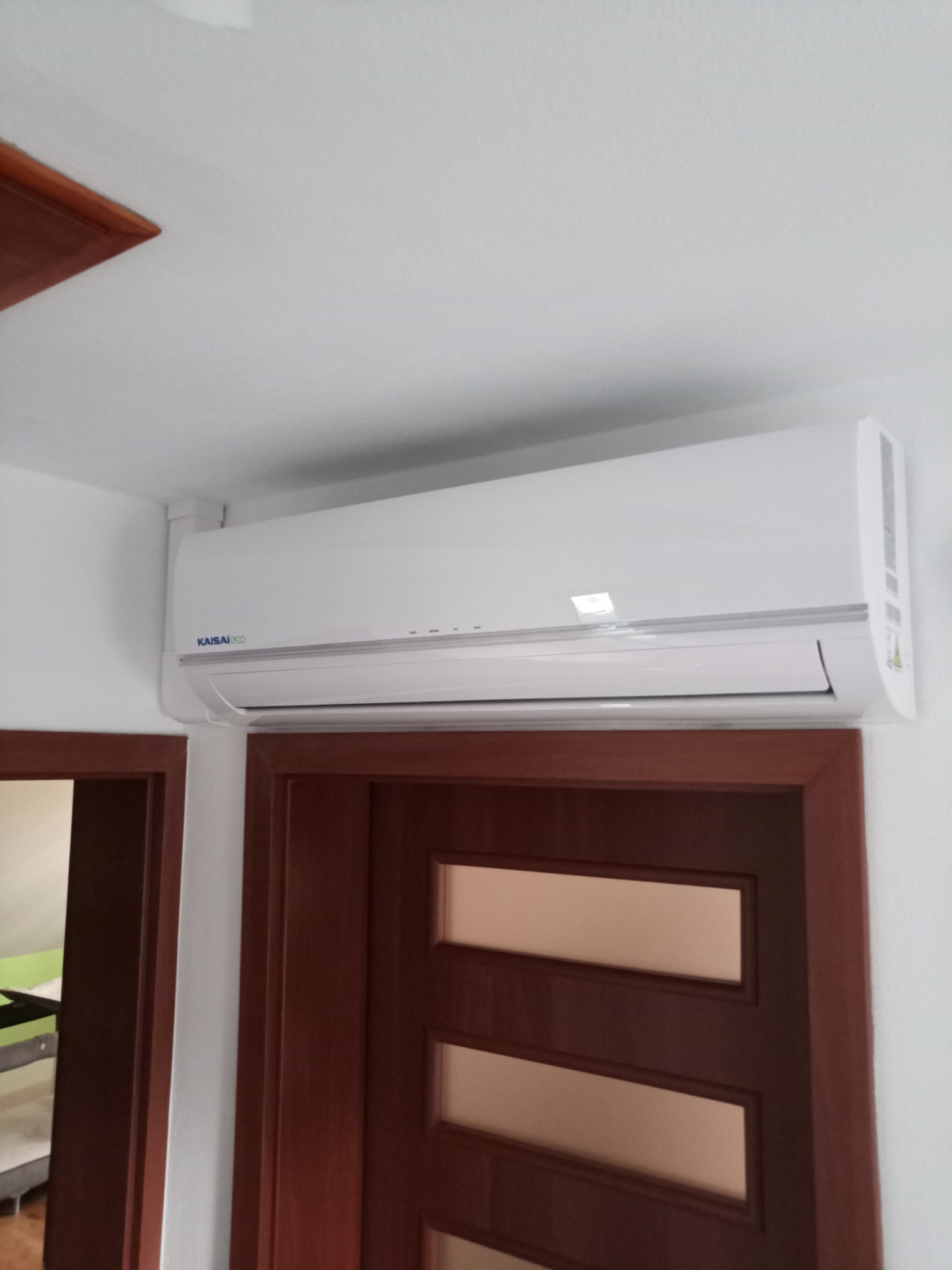Biały klimatyzator Kaisaieco zamontowany na ścianie nad brązowymi drzwiami z szybami w mieszkaniu.