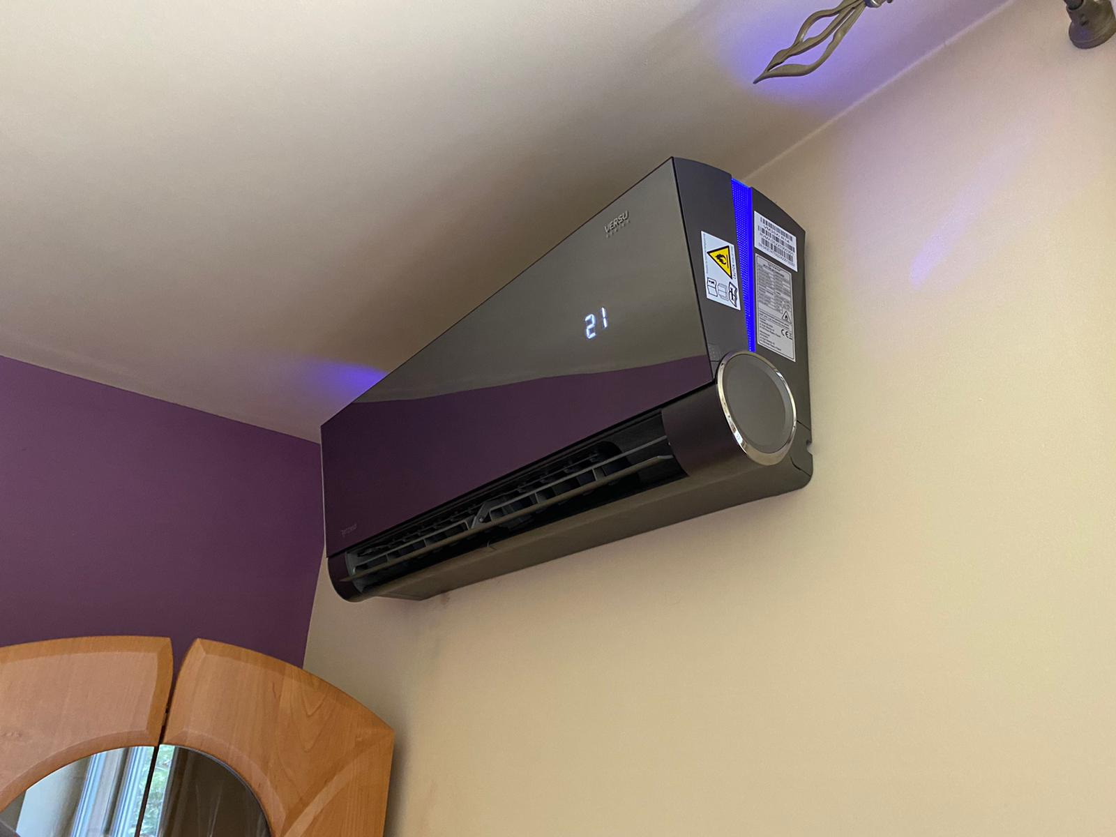 Czarny klimatyzator ścienny z wyświetlaczem ustawionym na 21 stopni, zamontowany w pokoju z fioletową ścianą i beżową ścianą, z widoczną dekoracją sufitową i fragmentem drewnianego mebla.