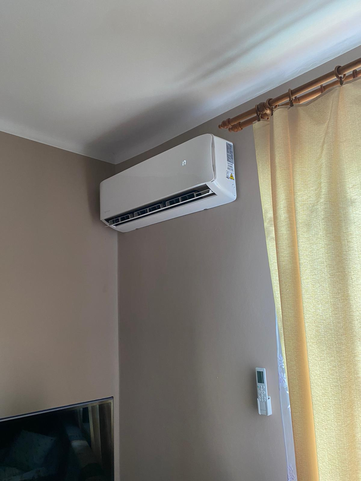 Biała jednostka klimatyzacyjna zamontowana na ścianie w pokoju z beżowymi ścianami, widoczny pilot na ścianie i fragment telewizora w rogu kadru.