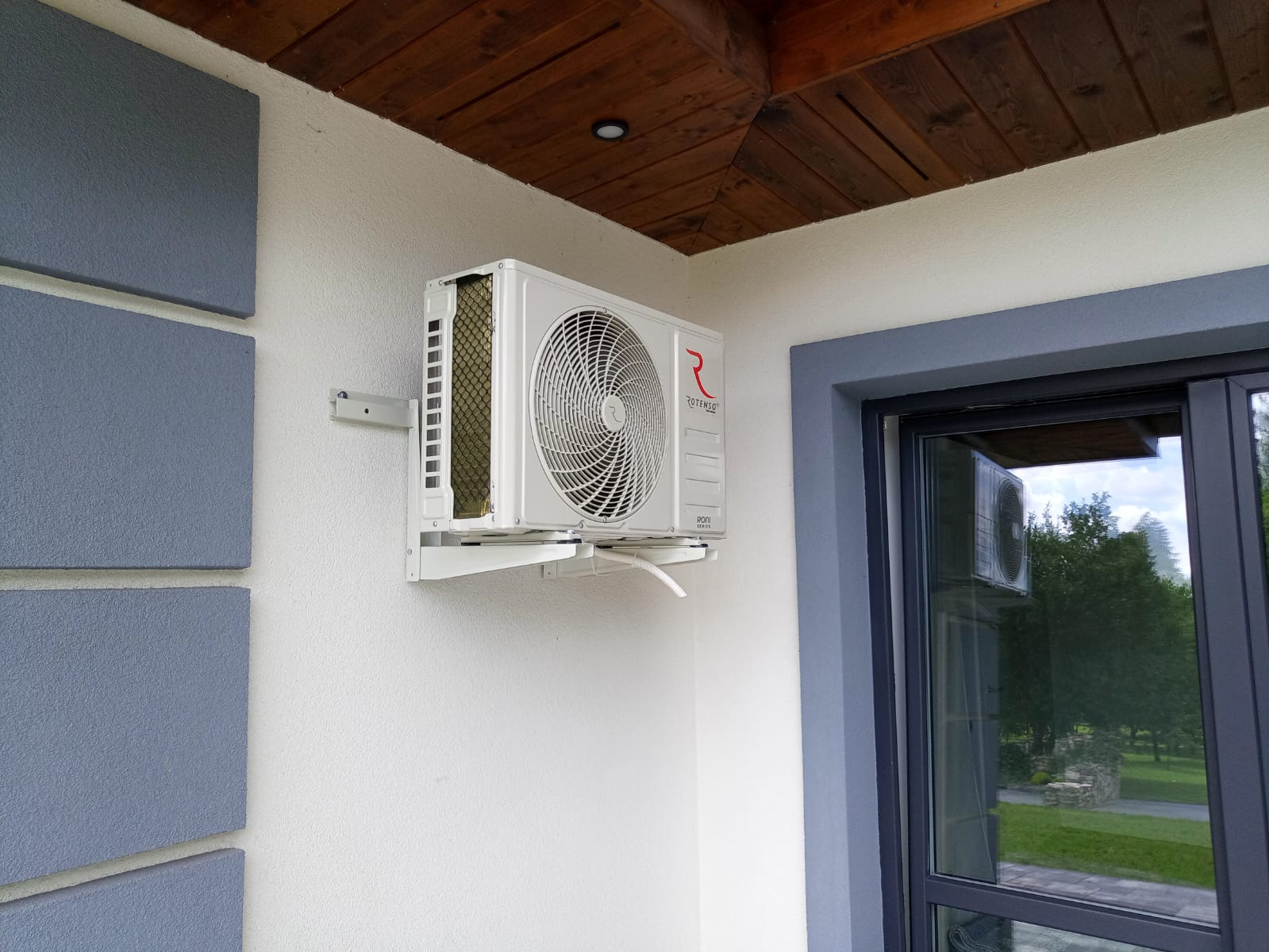 Biała jednostka zewnętrzna klimatyzacji Rotenso zamontowana na białej ścianie budynku z drewnianą podbitką dachową, widoczna obok okna z szarą ramą.