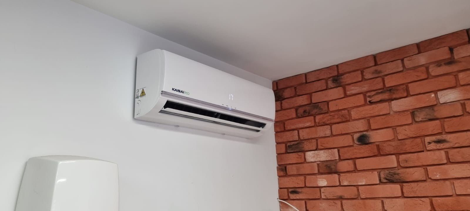Biała klimatyzacja Kaisai Eco zamontowana na białej ścianie, obok ściany z cegły dekoracyjnej w odcieniach czerwieni i brązu. Na wyświetlaczu klimatyzatora widoczna temperatura 17 stopni Celsjusza.