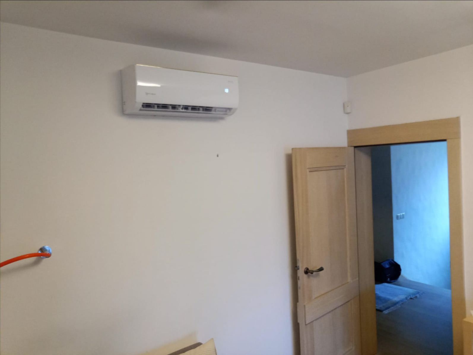 Biały klimatyzator ścienny zamontowany w jasnym pomieszczeniu, widoczny fragment instalacji z rurką w lewym dolnym rogu oraz otwarte drzwi do kolejnego pokoju.