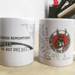 Dwa białe kubki z nadrukiem: jeden z danymi kontaktowymi firmy remontowej, drugi z logo firmy przedstawiającym muskularnego łosia w stroju roboczym. W tle widoczna maszyna do nadruków.
