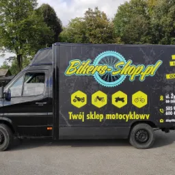 Czarny samoch&oacute;d dostawczy z reklamą sklepu motocyklowego Bikers-Shop.pl, z logo i danymi kontaktowymi, zaparkowany na tle drzew i zabudowań.