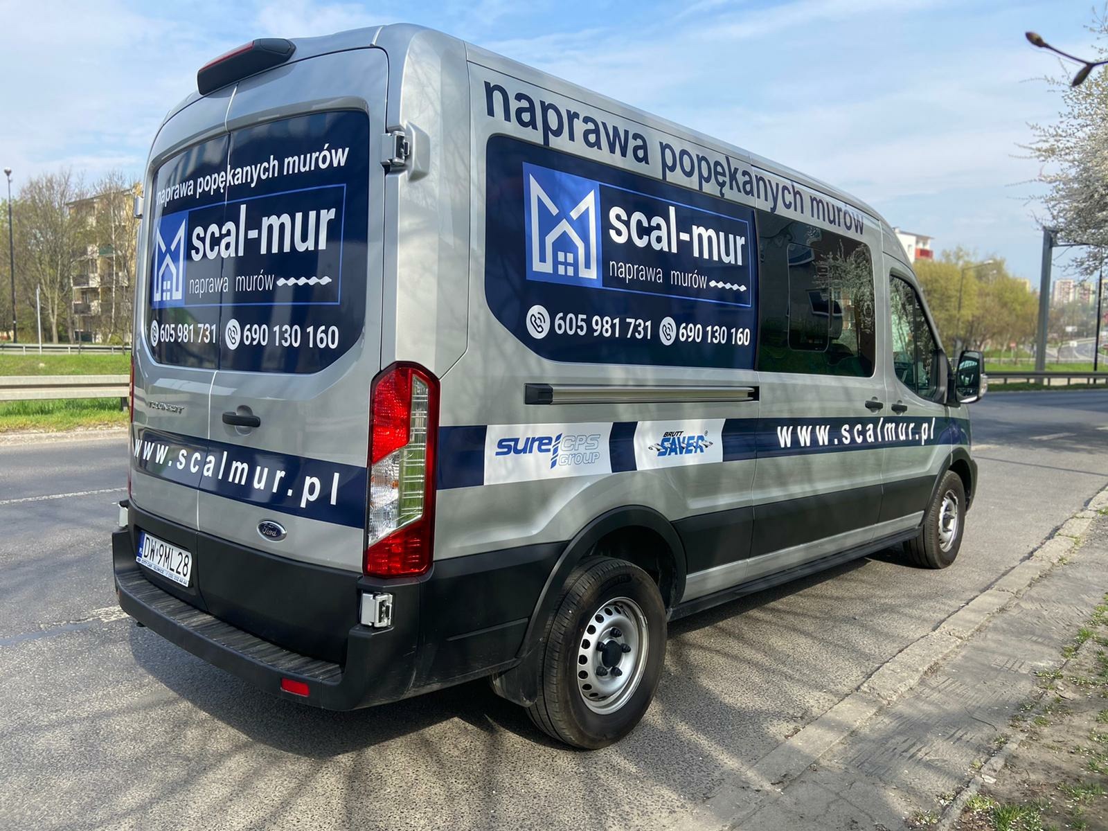 Srebrny samochód dostawczy z reklamą firmy 'scal-mur' zajmującej się naprawą popękanych murów, zaparkowany na ulicy w słoneczny dzień.