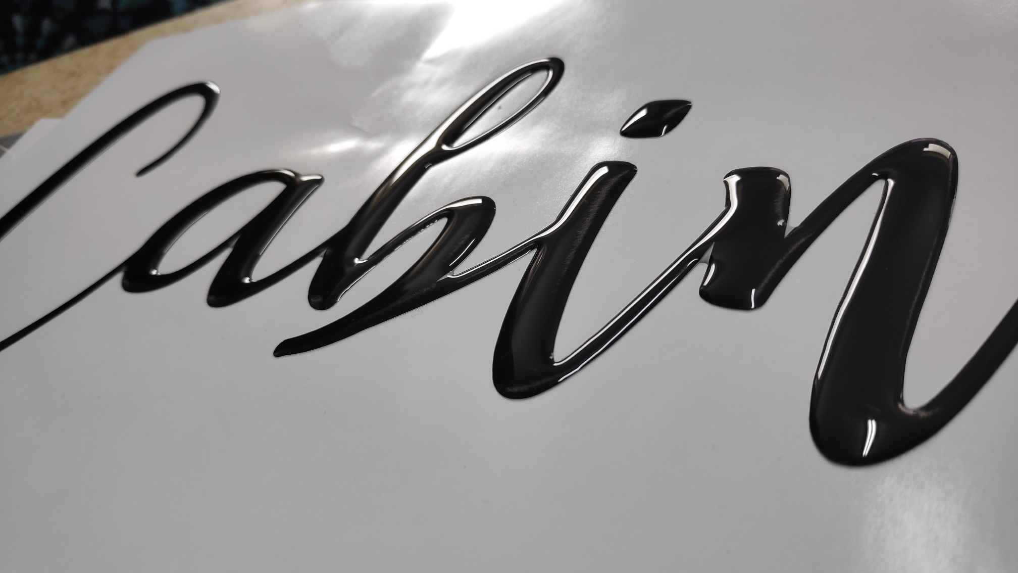 Błyszczący, czarny napis 'Cabin' wykonany techniką wylewania na gładkiej, białej powierzchni, widoczne refleksy światła.