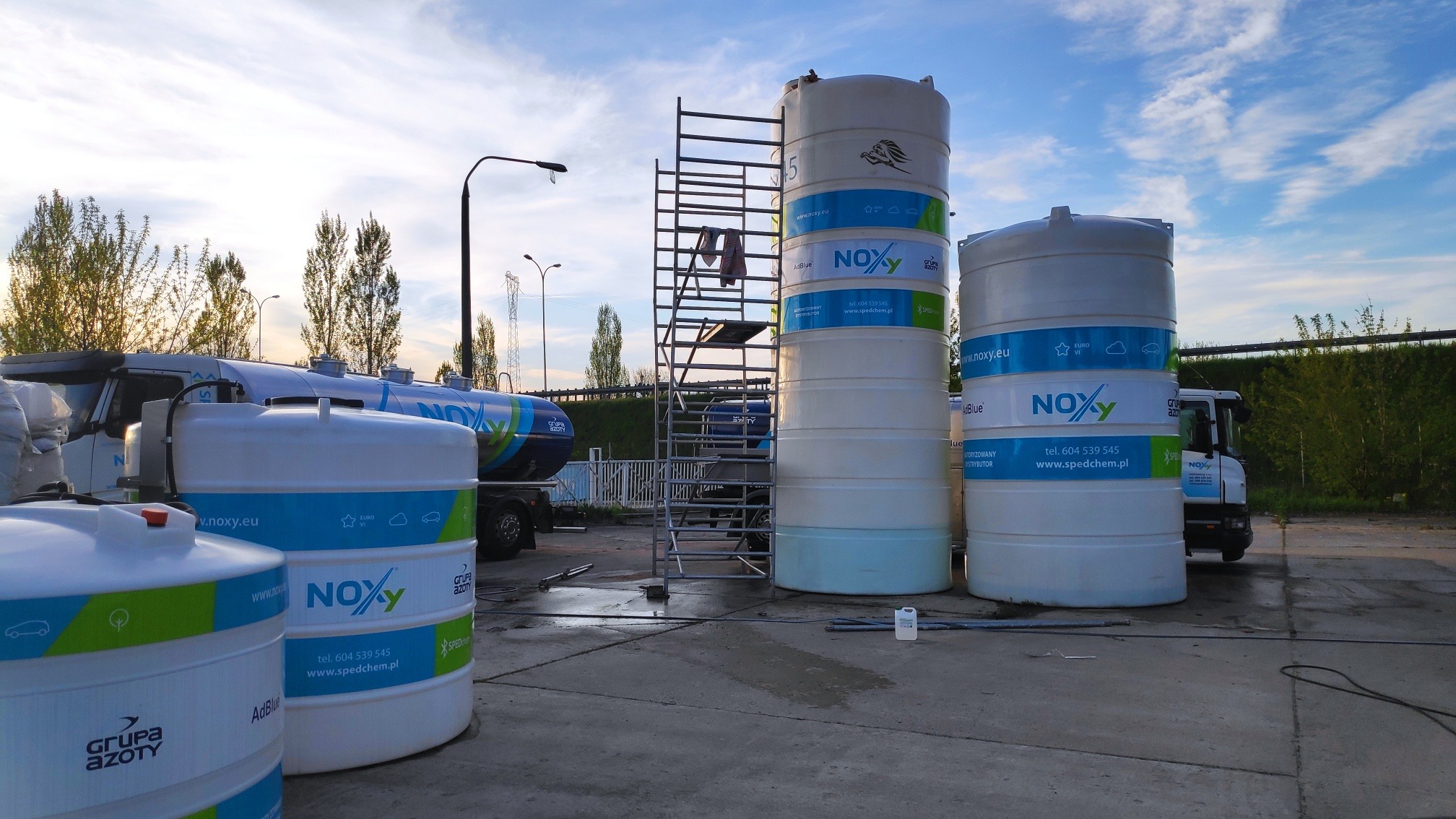 Białe zbiorniki z niebieskimi i zielonymi elementami graficznymi i logo NOXY na tle trucka i metalowych schodów, na zewnątrz, w pochmurny dzień.