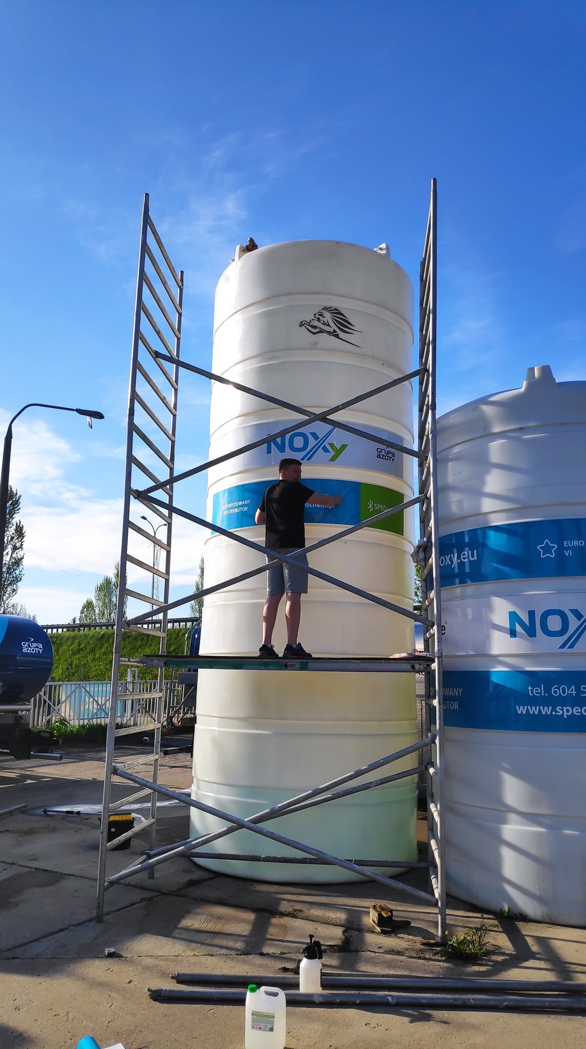 Pracownik na rusztowaniu nakleja folię z logo i danymi kontaktowymi na biały zbiornik NOXY Grupy Azoty, widoczne dodatkowe zbiorniki i butelki z płynami.