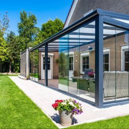 GREMAG Sp. z o.o. Salon Fachmann Polkowice - Szklana zabudowa tarasu z metalową konstrukcją, widok z zewnątrz na tle zielonego trawnika i budynku z cegły. Wnętrze z meblami ogrodowymi.