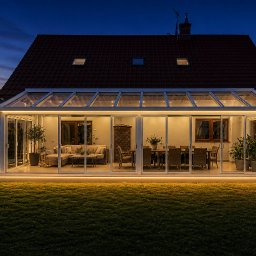 Elitegarden - Oświetlona, przeszklona zabudowa tarasowa z metalową konstrukcją, widoczna z zewnątrz w nocy. Wewnątrz meble wypoczynkowe i jadalniane.