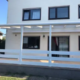 Biała metalowa konstrukcja zadaszenia tarasu z barierkami, przylegająca do jasnego budynku z ciemnymi oknami, widok z zewnątrz w słoneczny dzień.