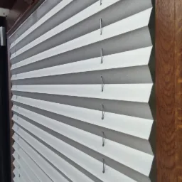 Szczegółowe ujęcie białych, plisowanych osłon okiennych z widocznymi sznurkami, zamontowanych w oknie z ciemnobrązową ramą.