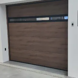 Segmentowa brama garażowa w kolorze drewna z czarnymi detalami, widoczna wewnątrz pomieszczenia na tle białych ścian.