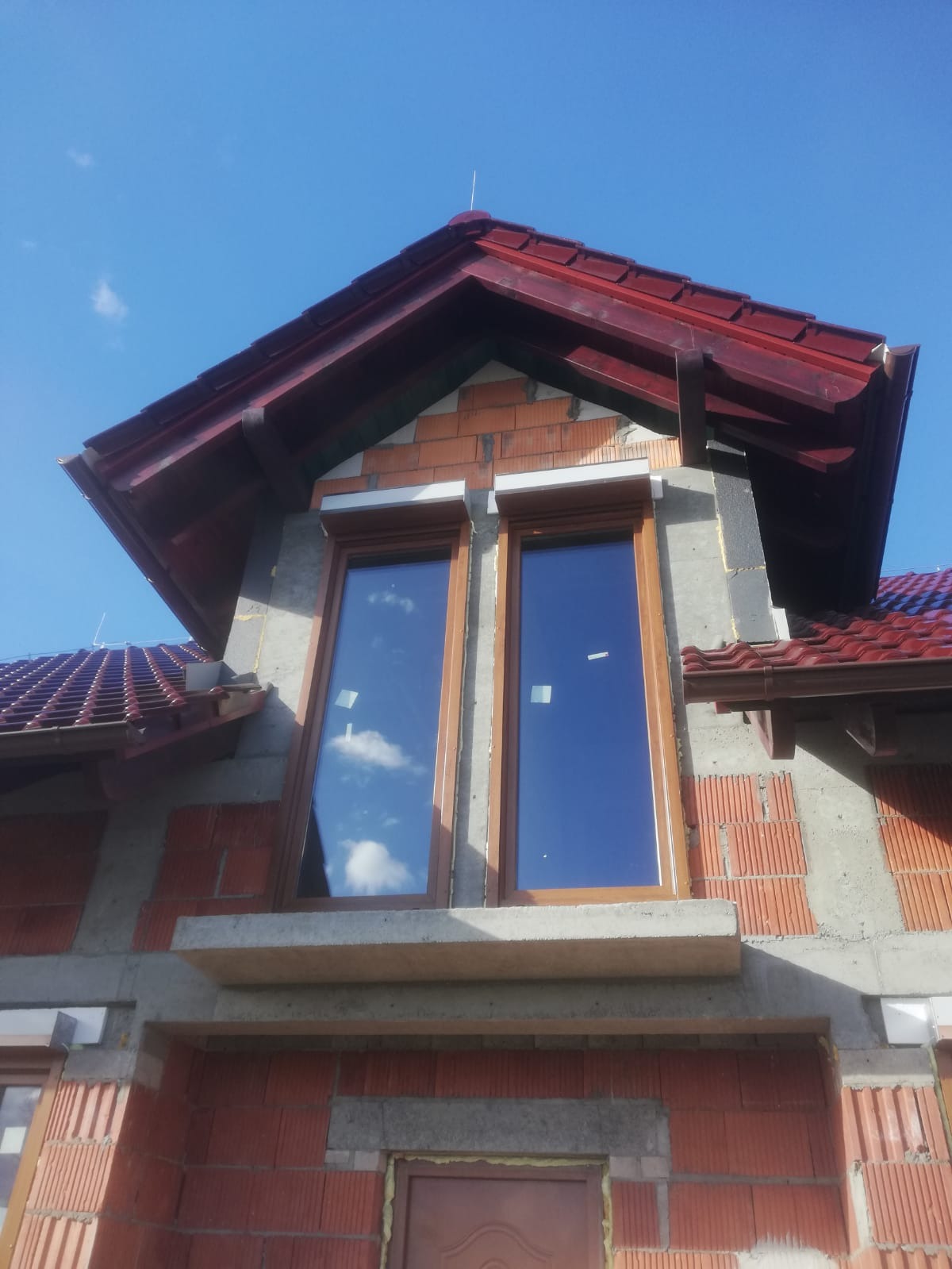 Montaż okien z brązowymi ramami w surowym stanie budynku z widocznymi pustakami ceramicznymi i częściowo otynkowanymi ścianami, odbicie błękitnego nieba z chmurami w szybach.