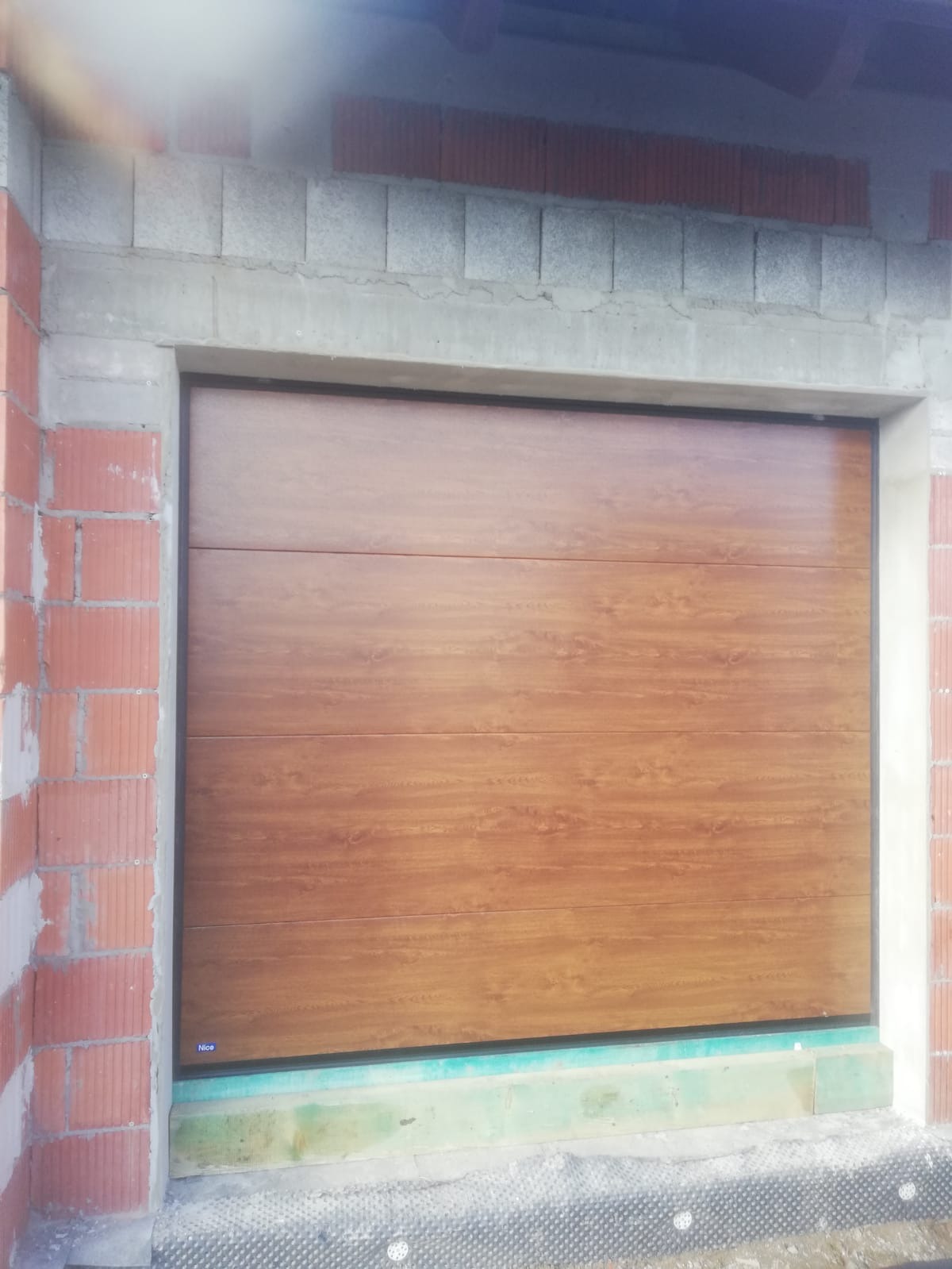Panelowa brama garażowa w odcieniach brązu, zamontowana w surowym otworze z cegły i betonu, widoczna z bliska podczas montażu.