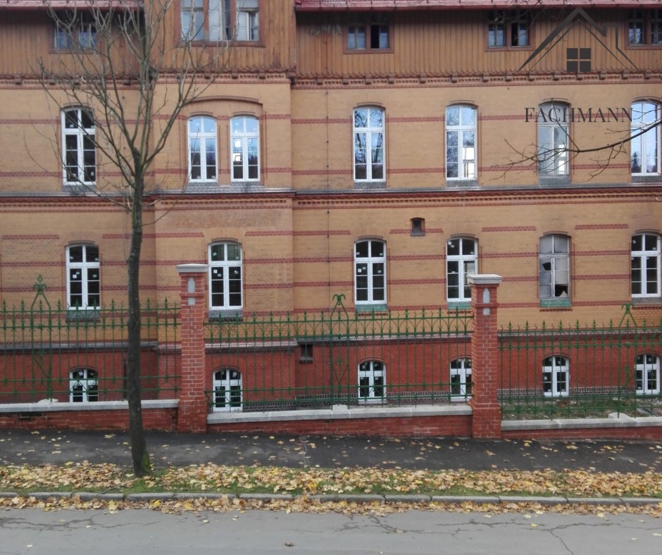 Fasada budynku z cegły z białymi oknami, zielonym ogrodzeniem i liśćmi na chodniku; jedno okno uszkodzone.