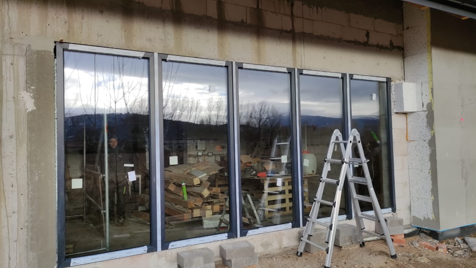 Montaż okien z szarymi ramami w surowym budynku, widoczne drabiny, materiały budowlane i odbicie pracownika w szybie.