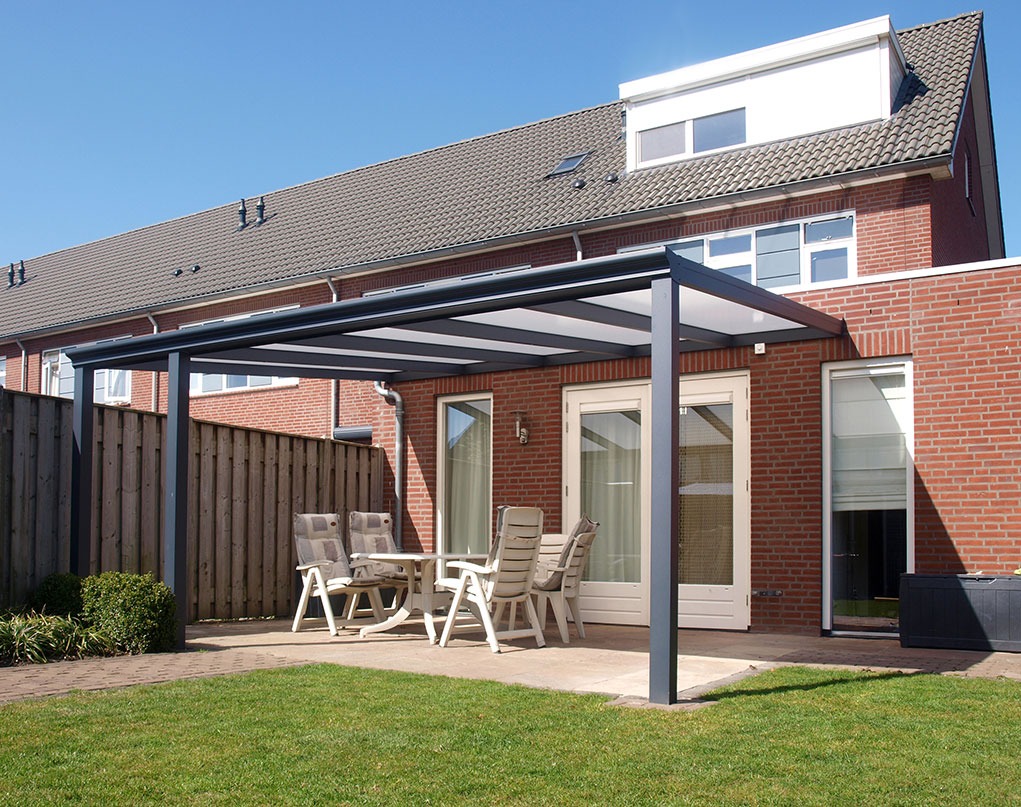 Nowoczesna pergola metalowa z przezroczystym zadaszeniem, ustawiona na tarasie przy domu z cegły, z widocznymi krzesłami i stołem. Słoneczny dzień.