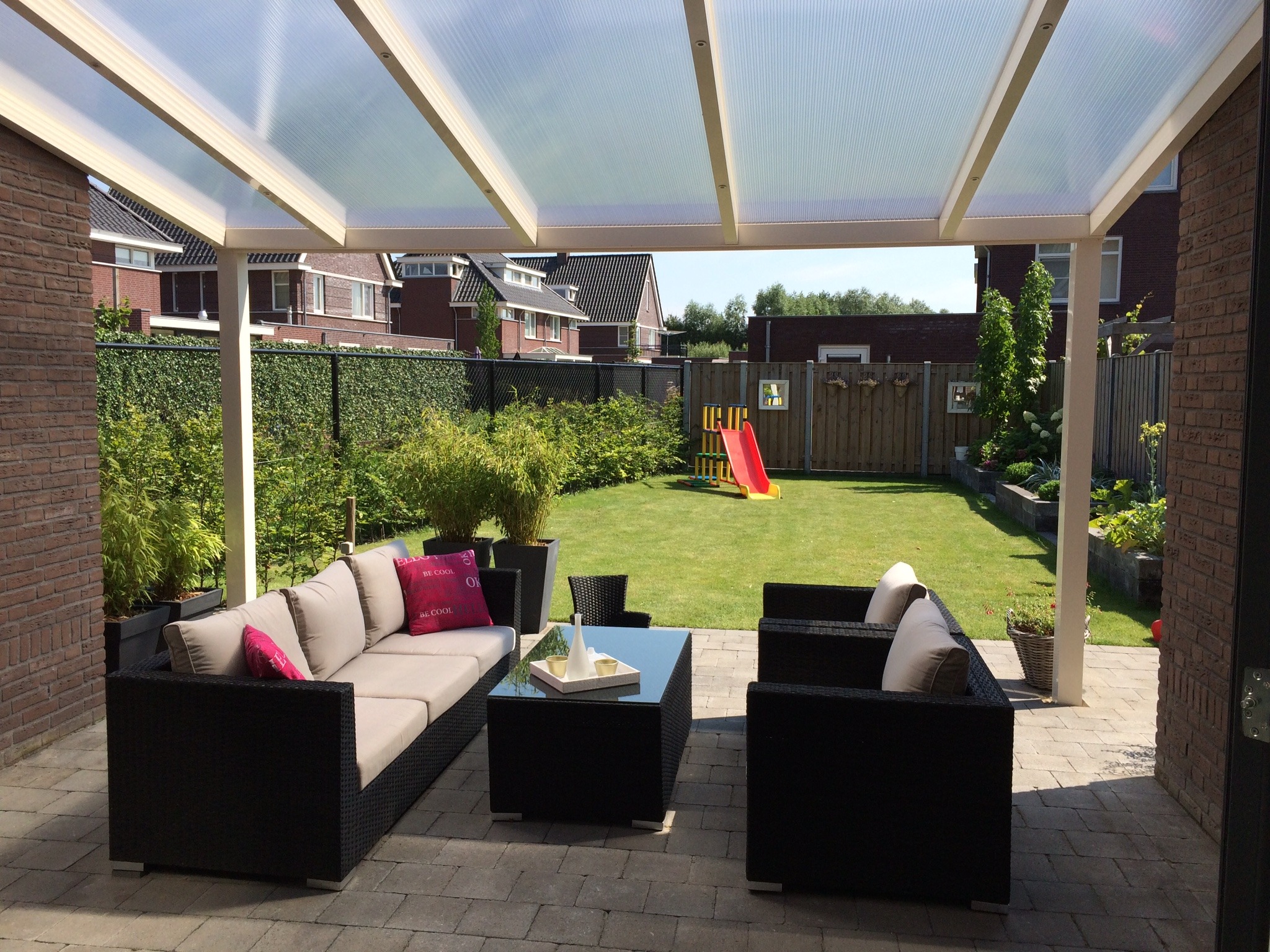 Nowoczesna pergola metalowa z przezroczystym zadaszeniem, pod którą znajdują się meble ogrodowe, tworząc strefę relaksu na tarasie z widokiem na zadbany ogród.