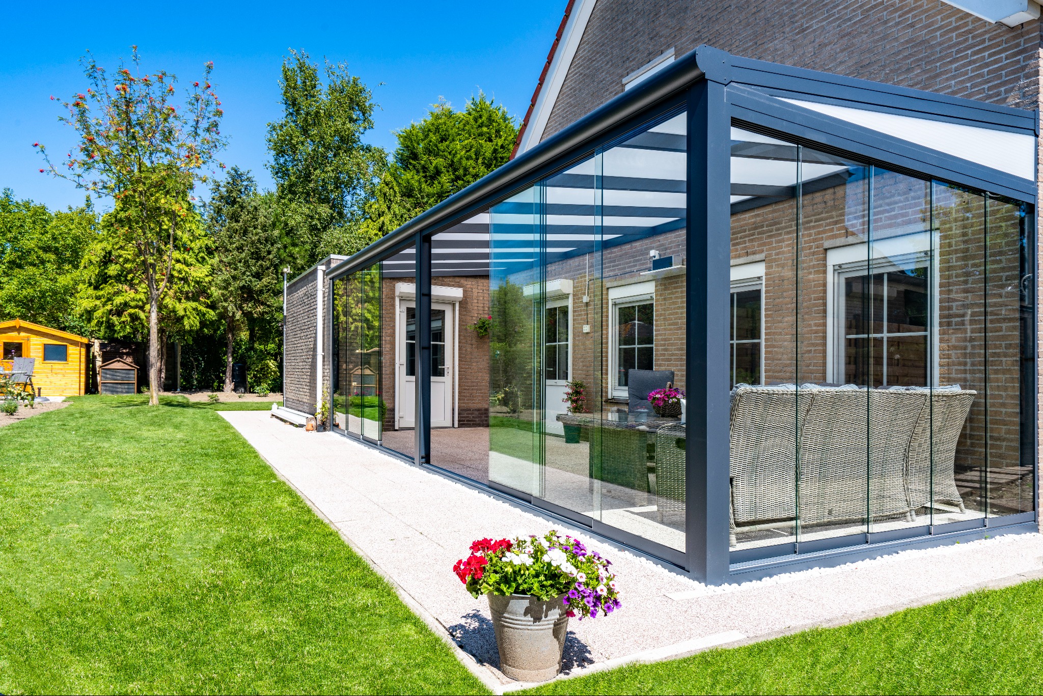 Szklana zabudowa tarasu z metalową konstrukcją, widok z zewnątrz na tle zielonego trawnika i budynku z cegły. Wnętrze z meblami ogrodowymi.