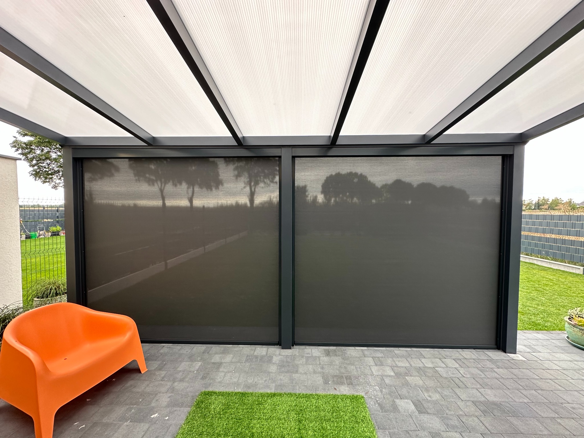 Zadaszona przestrzeń z szarymi roletami bocznymi, pomarańczowym krzesłem i sztuczną trawą. Widok na ogród przez rolety.
