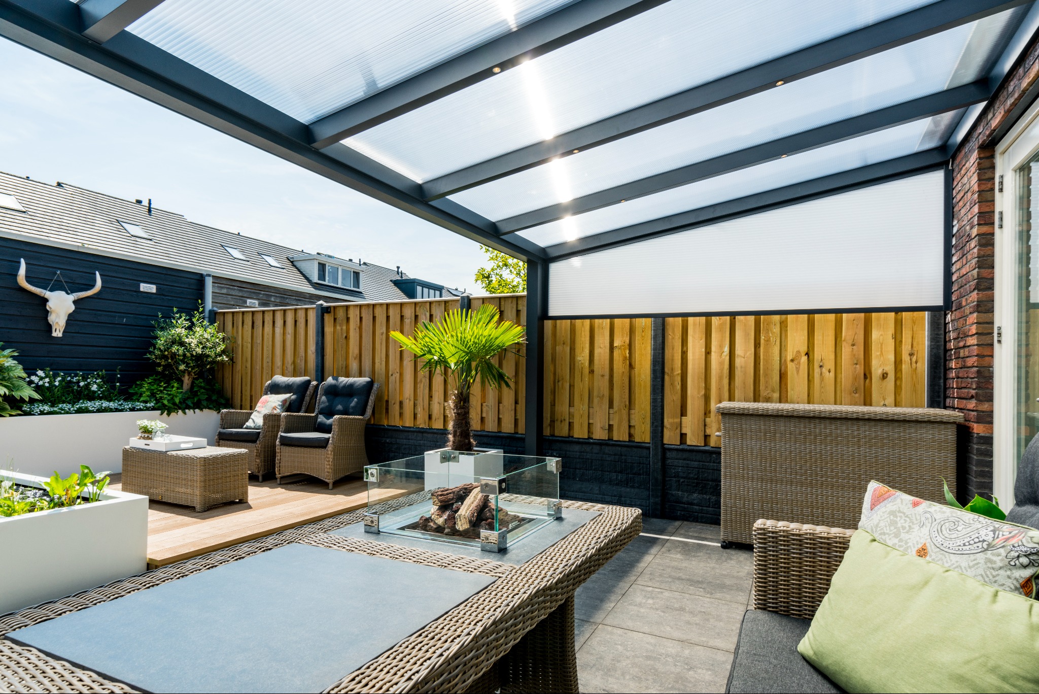 Zadaszony taras z metalową pergolą, rattanowymi meblami i paleniskiem. Widoczne drewniane ogrodzenie i dekoracje, w tym czaszka byka na ścianie.