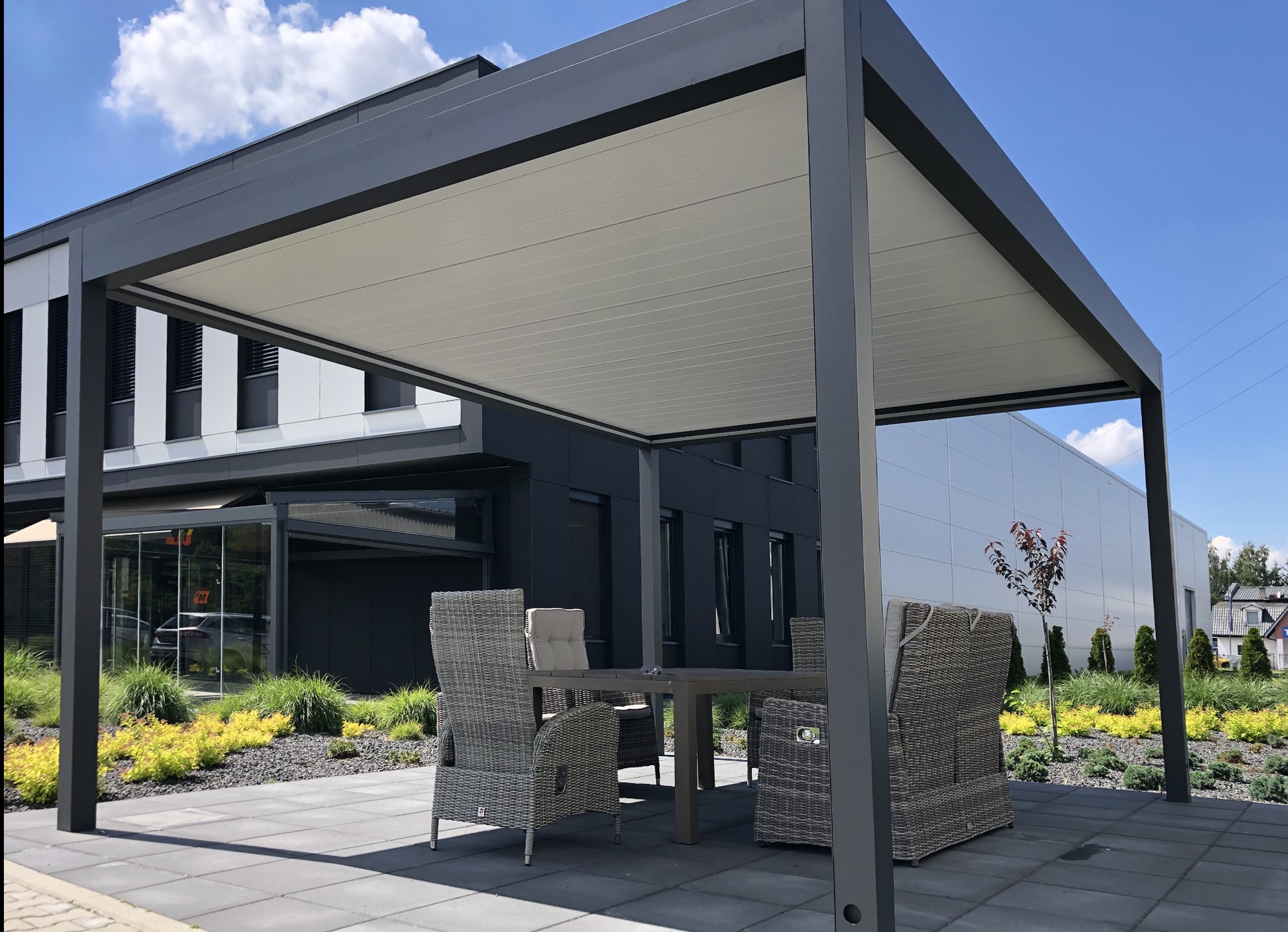 Nowoczesna, szara pergola metalowa z płaskim dachem, ustawiona na tarasie z szarych płyt. Wewnątrz zestaw mebli ogrodowych z rattanu. Tło stanowi budynek z elementami szkła.