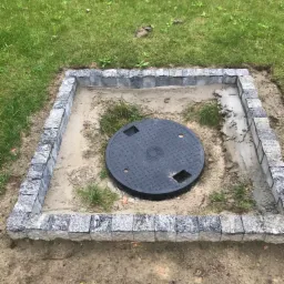 Obrzeże z kostki granitowej wokół studzienki kanalizacyjnej na trawniku, widoczne ślady zaprawy.
