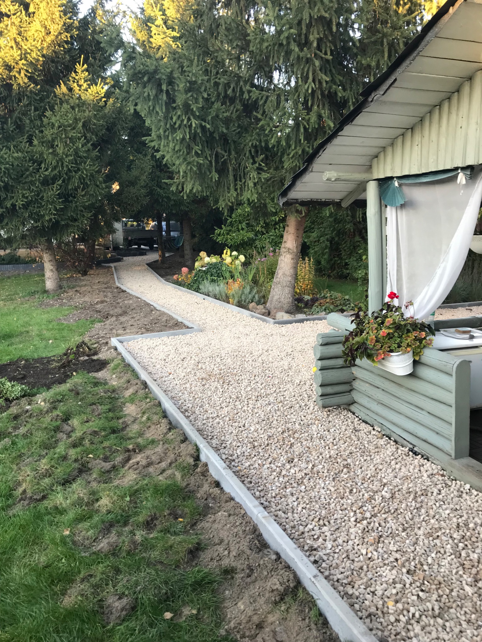 Kątowe ujęcie ścieżki z jasnego żwiru, obramowanej betonowymi obrzeżami, prowadzącej przez trawnik w kierunku domku ogrodowego z dekoracyjną zasłoną, w tle widoczne drzewa i pojazd.