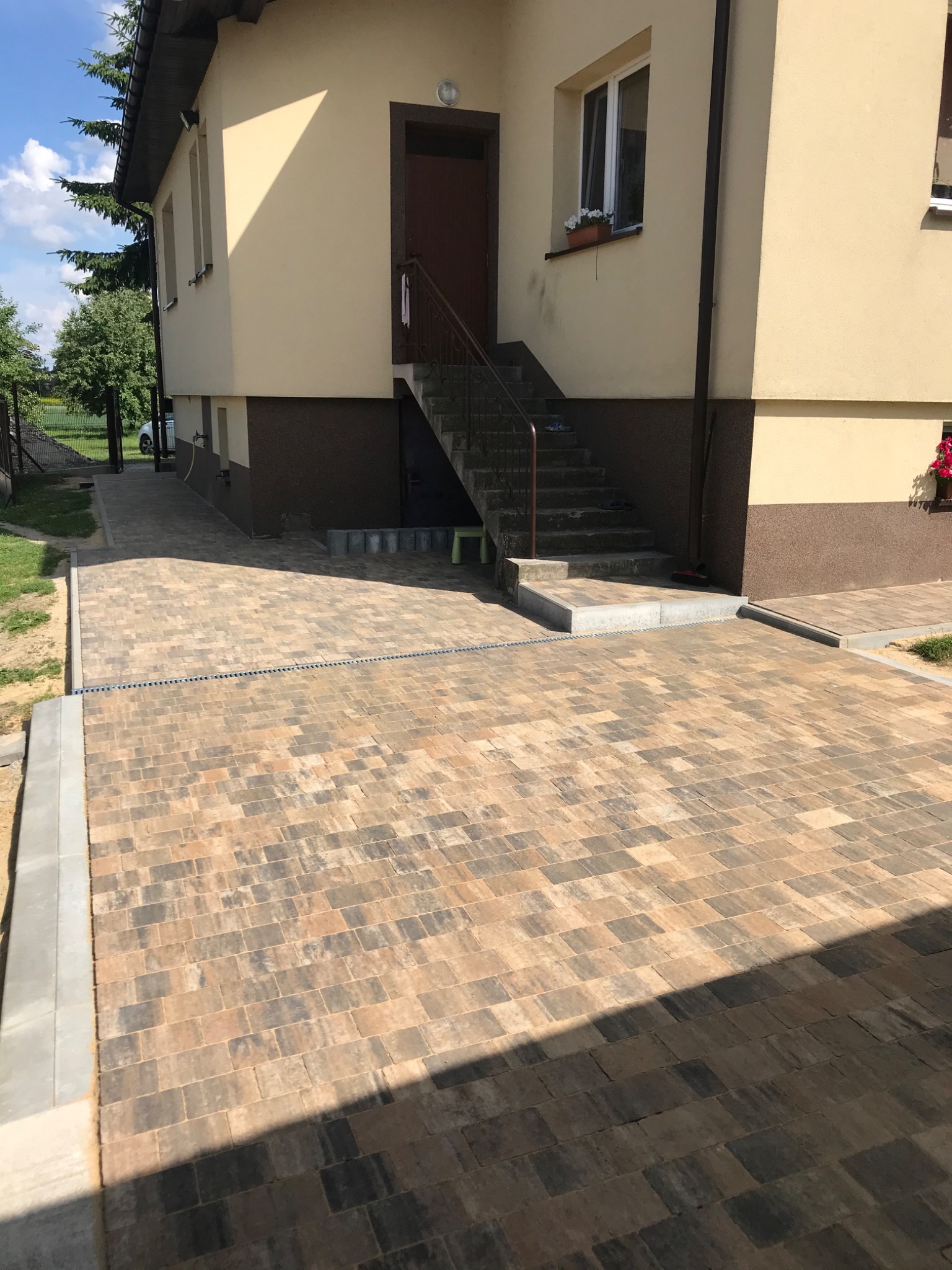 Nowo ułożony, wzorzysty podjazd z kostki brukowej w odcieniach brązu i szarości, z widocznym odpływem liniowym, prowadzący do schodów wejściowych domu z jasną elewacją.