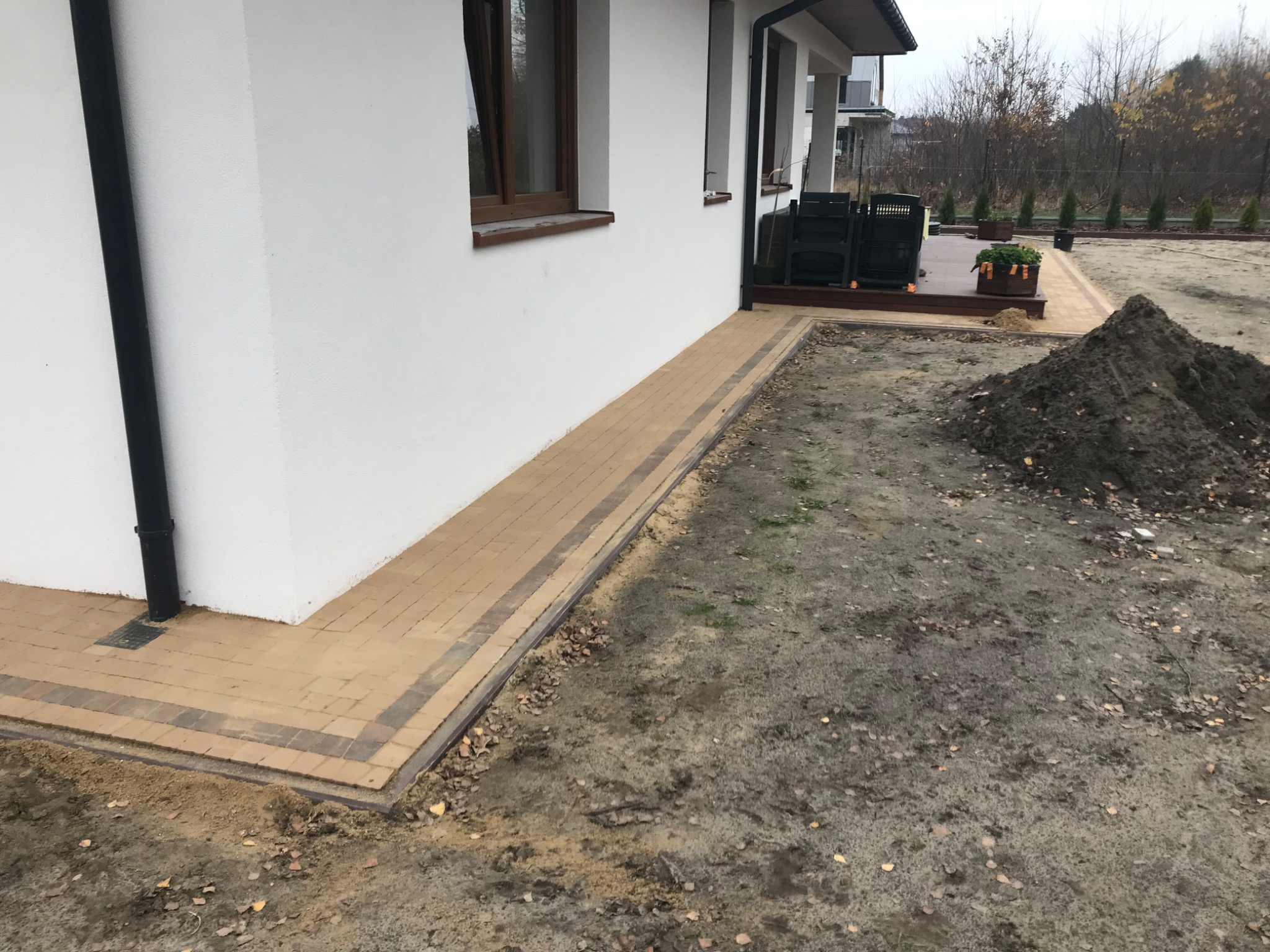 Ścieżka z kostki brukowej obok białego domu w Brwinowie, widoczny fragment trawnika i pryzma ziemi, prace w toku.