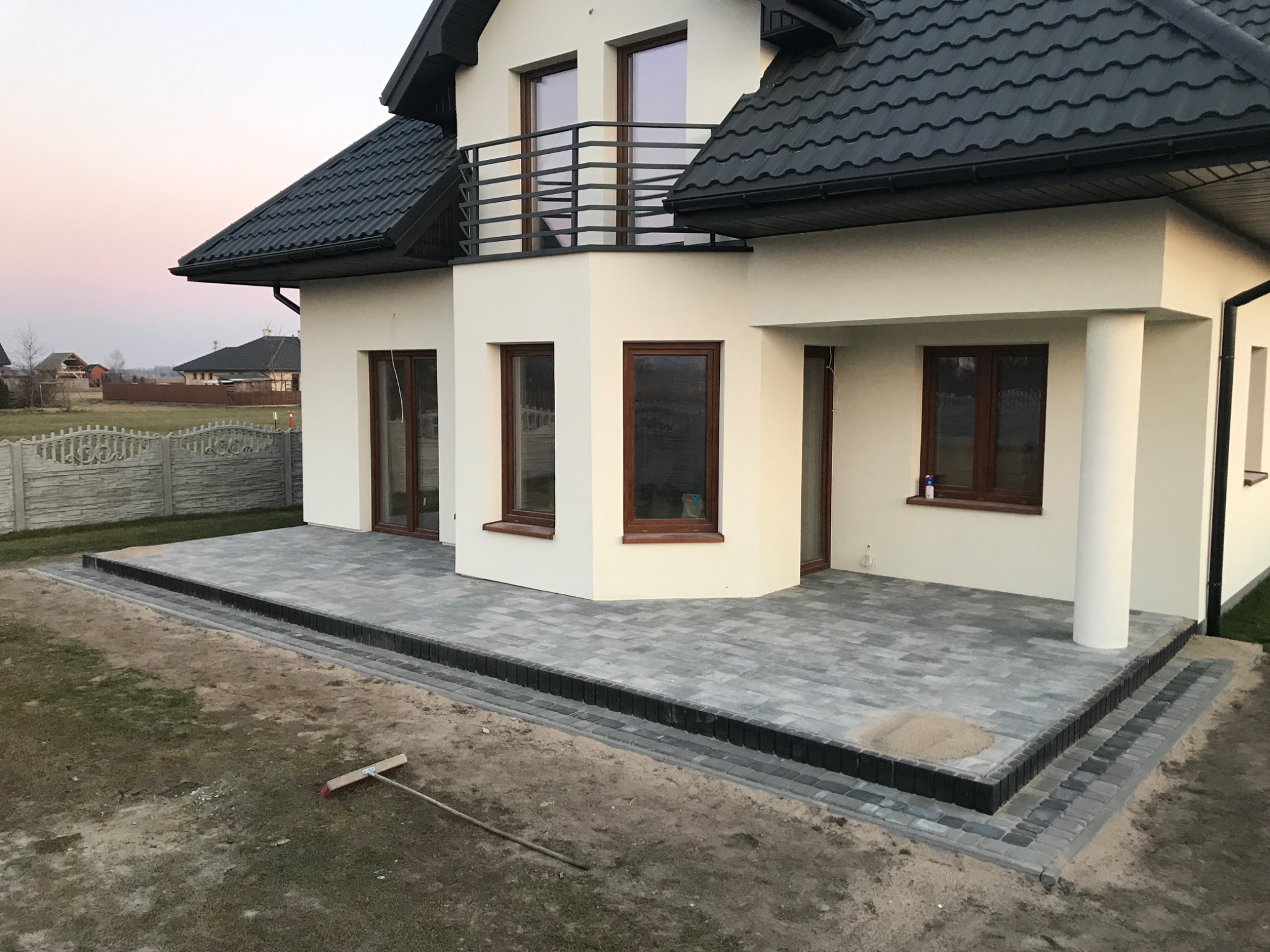 Nowoczesny taras z szarej kostki brukowej przy jasnym domu z czarnym dachem; widoczna szczotka brukarska na ziemi.