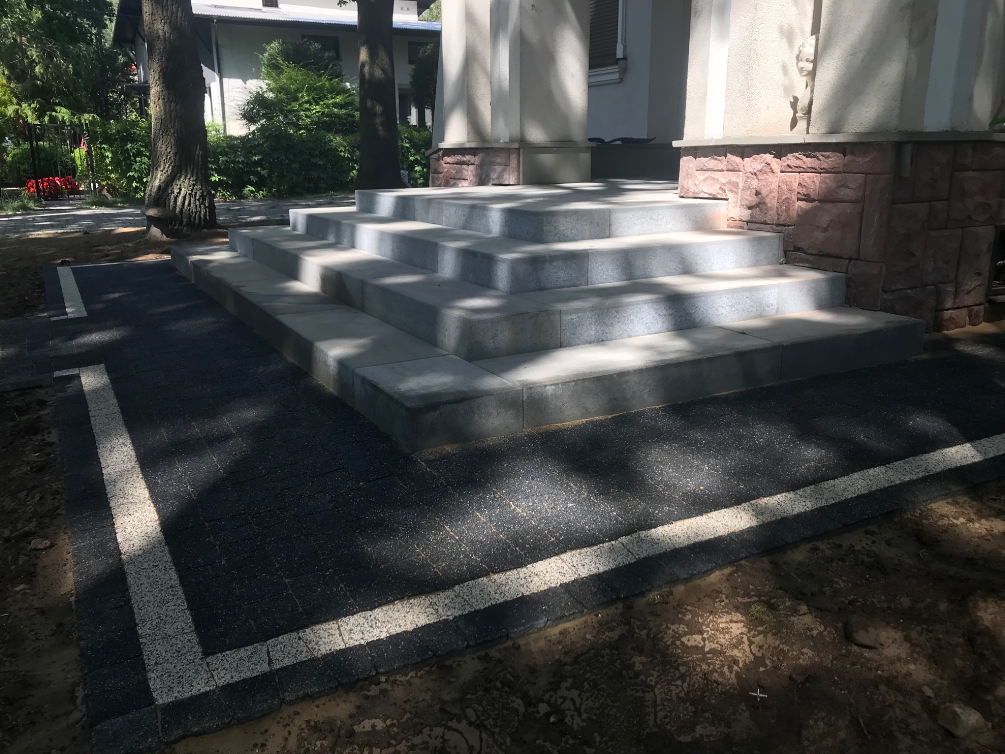 Nowe schody wejściowe z szarej kostki prowadzące do budynku, obrzeża z ciemnej kostki z jasnymi liniami, widoczne elementy architektury budynku.