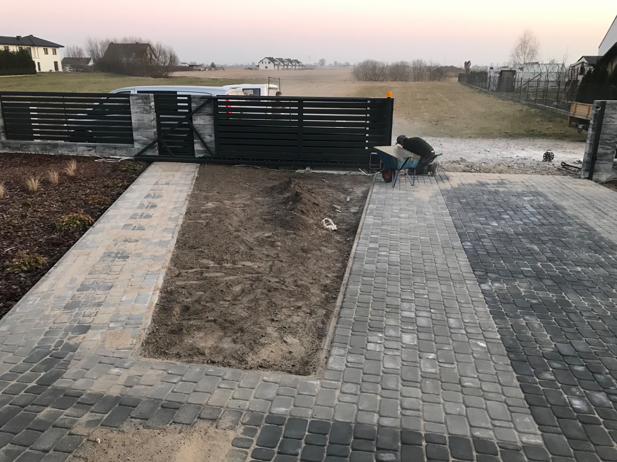 Pracownik na kolanach wyrównuje podłoże pod kostkę w wykopie otoczonym świeżo ułożoną, szarą kostką brukową w dwóch odcieniach, w tle brama wjazdowa i zabudowa jednorodzinna.