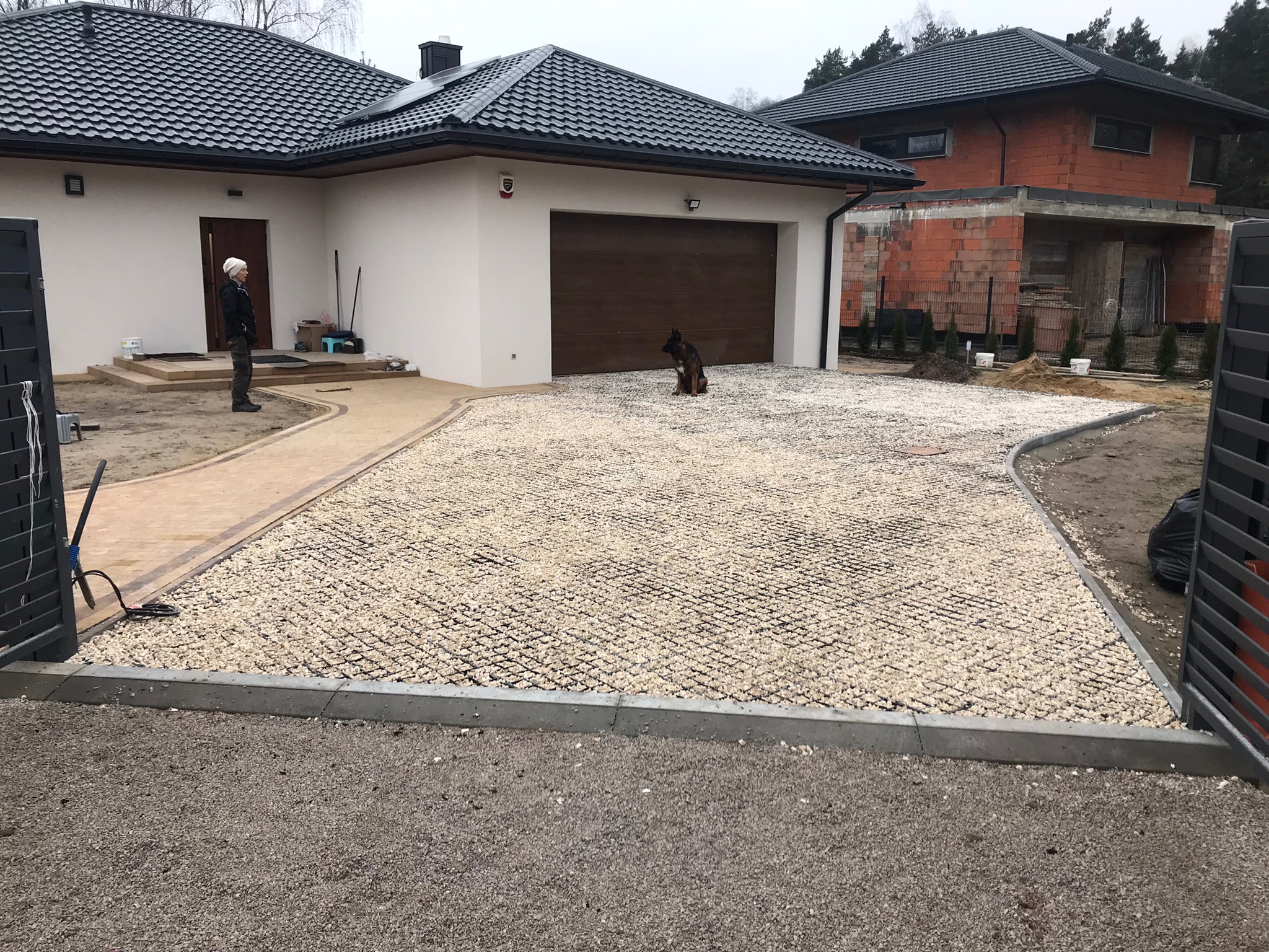 Niedokończony podjazd z białym żwirem stabilizowanym kratką, obrzeża z szarej kostki, w tle dom z garażem i budynek w trakcie budowy, pies siedzący na żwirze, pracownik w oddali.