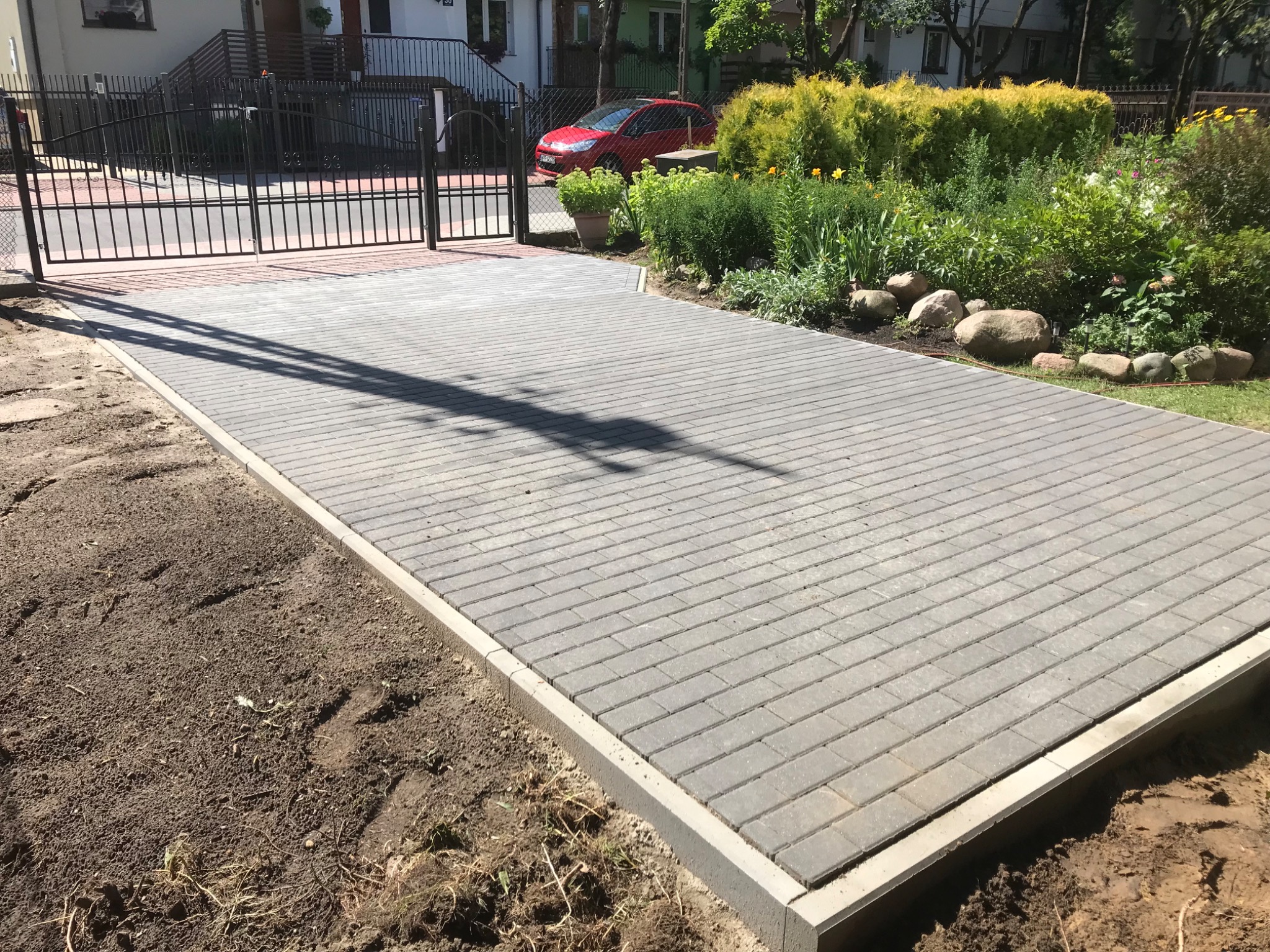 Ułożona szara kostka brukowa na podjeździe, widoczny fragment metalowej bramy w tle oraz rabata z roślinami i kamieniami.