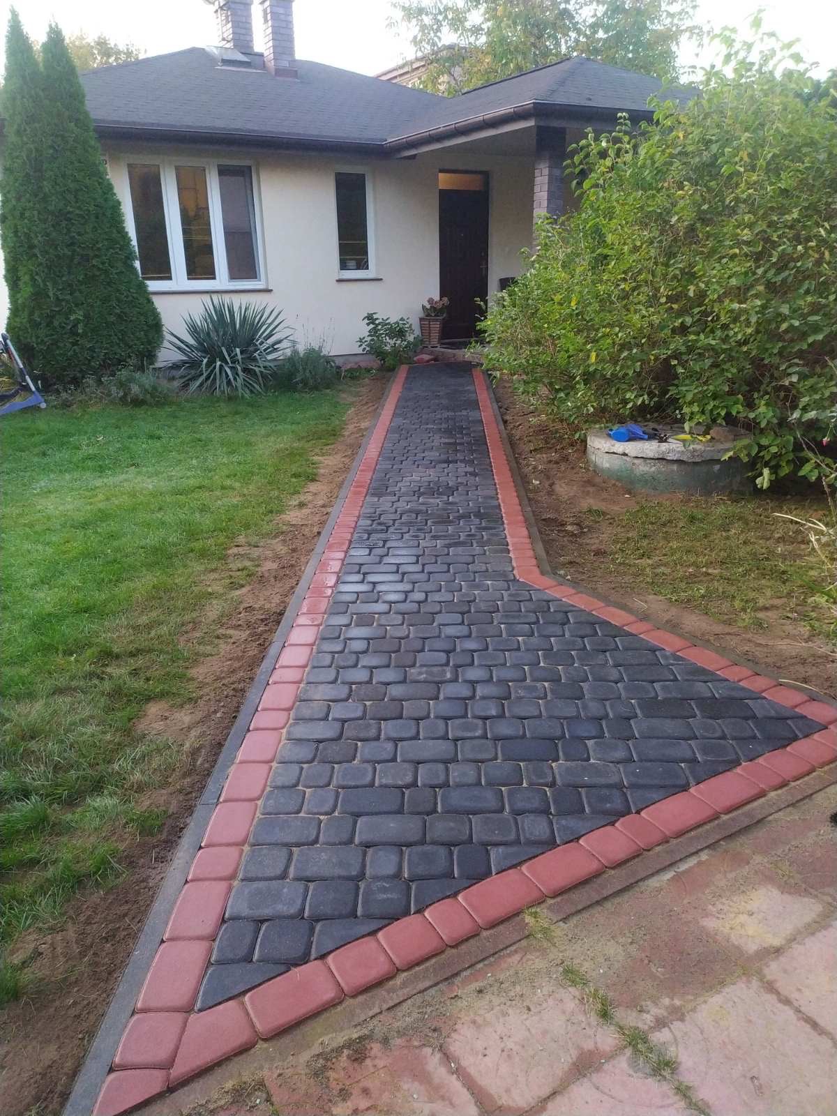 Ścieżka z ciemnoszarej kostki brukowej z obramowaniem z czerwonej kostki prowadząca do wejścia domu z ogrodem.