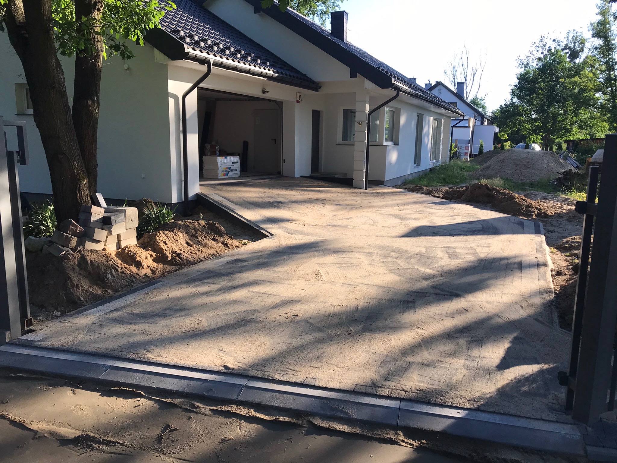 Ułożona kostka brukowa na podjeździe do garażu, widoczne obrzeża, w tle biały dom z czarnym dachem, paleta kostki brukowej w garażu, prace w toku.