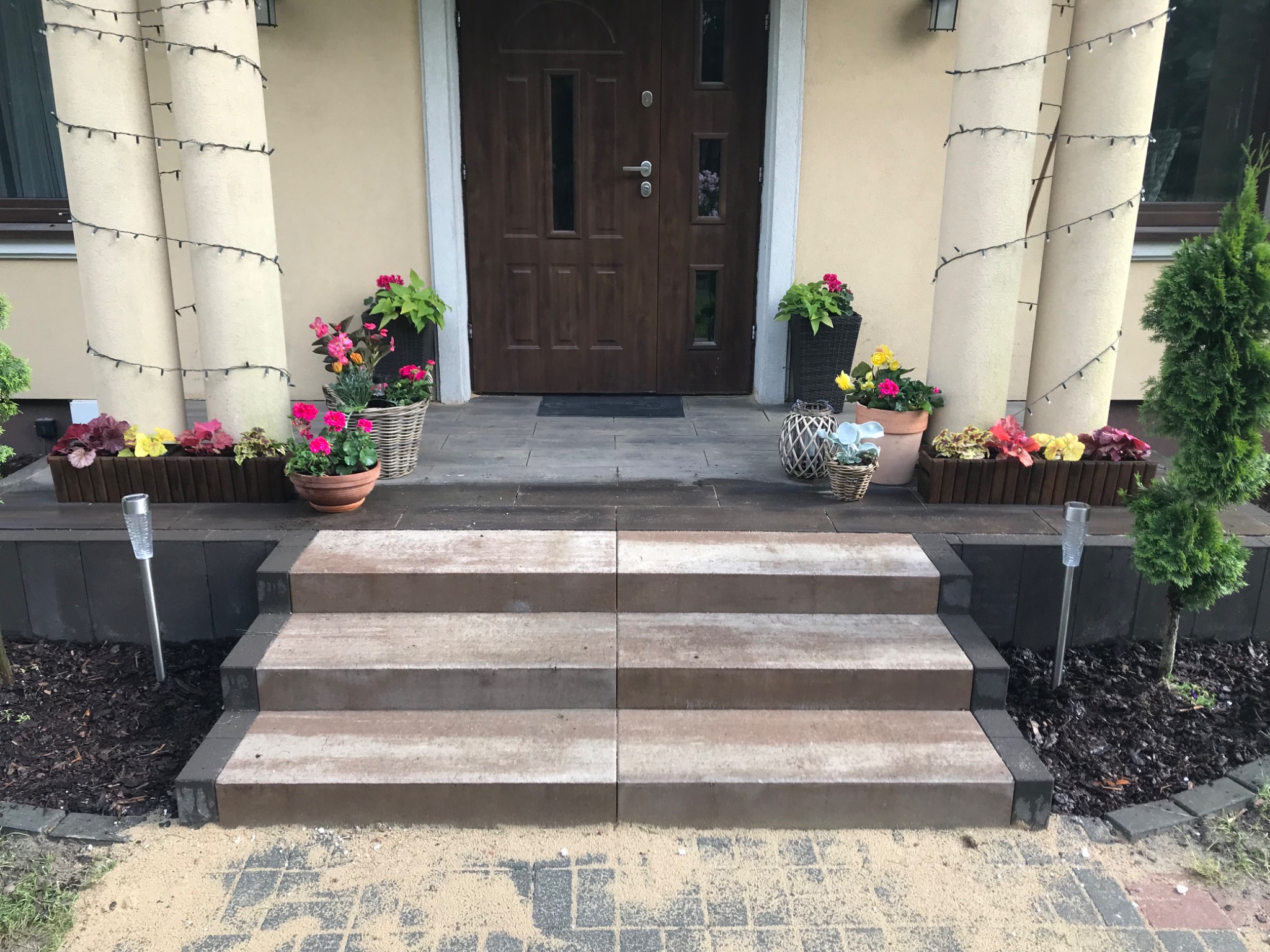 Schody wejściowe z kostki brukowej prowadzące do drzwi domu, ozdobione kwiatami w donicach i lampkami na kolumnach.