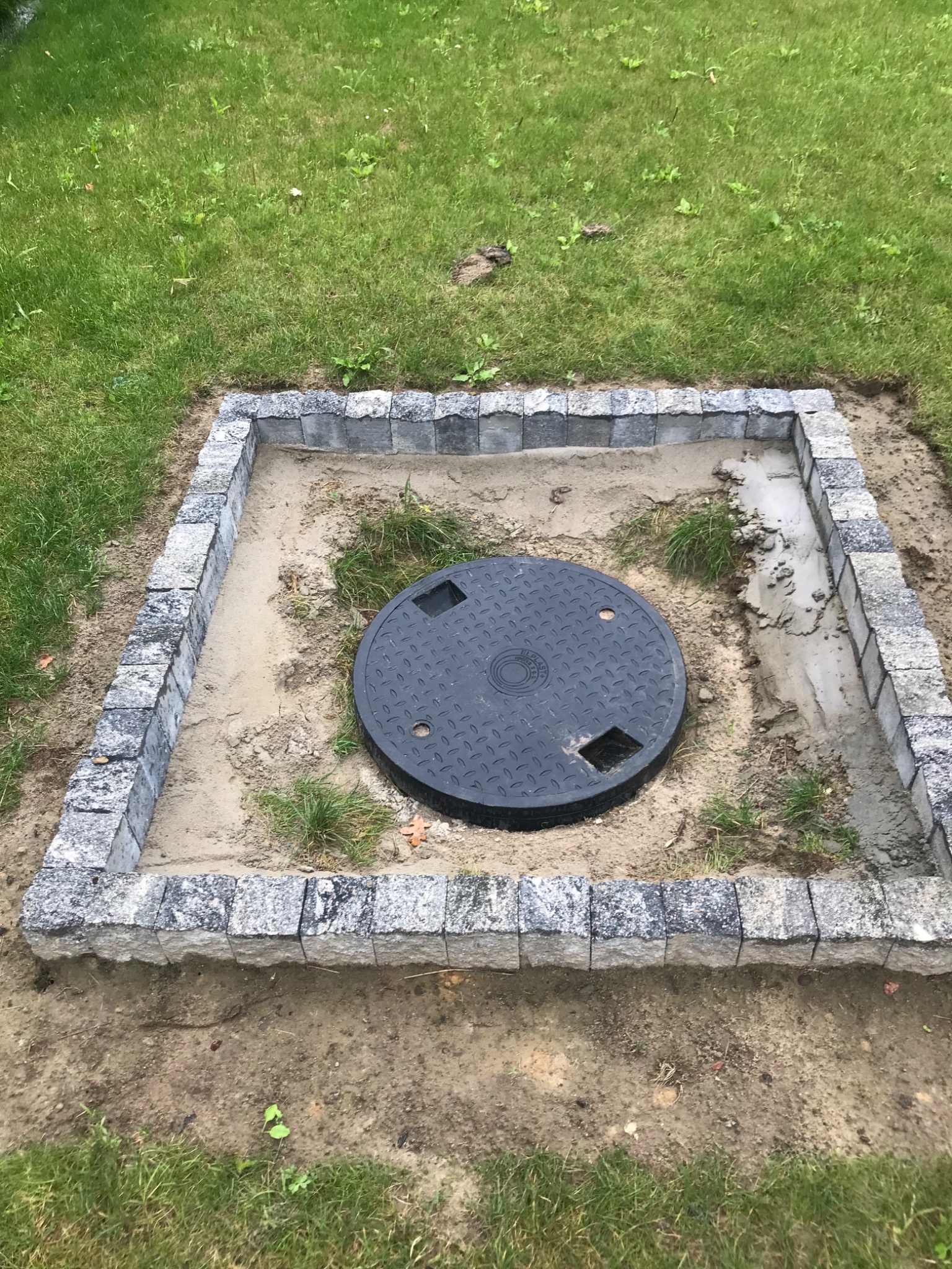 Obrzeże z kostki granitowej wokół studzienki kanalizacyjnej na trawniku, widoczne ślady zaprawy.