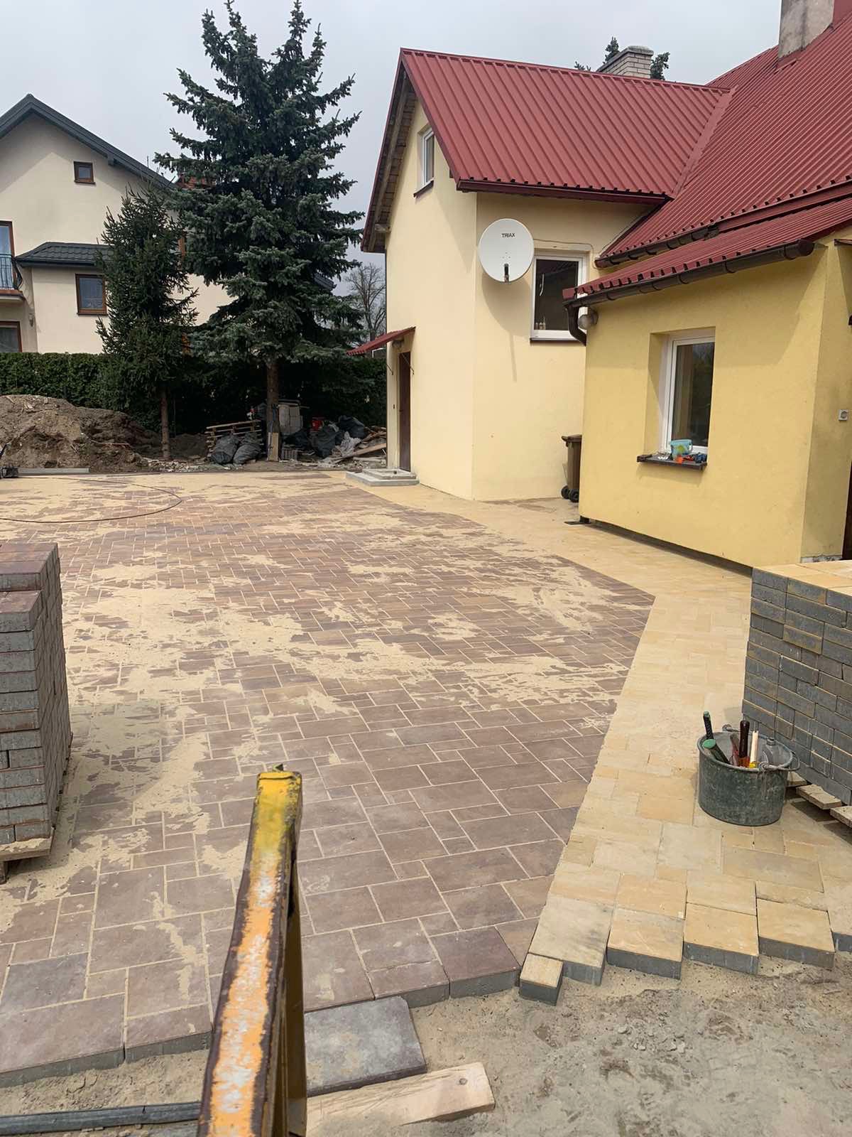 Układanie kostki brukowej na posesji: widok na rozległy plac z częściowo ułożoną, brązową kostką brukową, obok stos kostki, wiadro z narzędziami i żółty kozioł roboczy, w tle żółty dom z czerwonym...