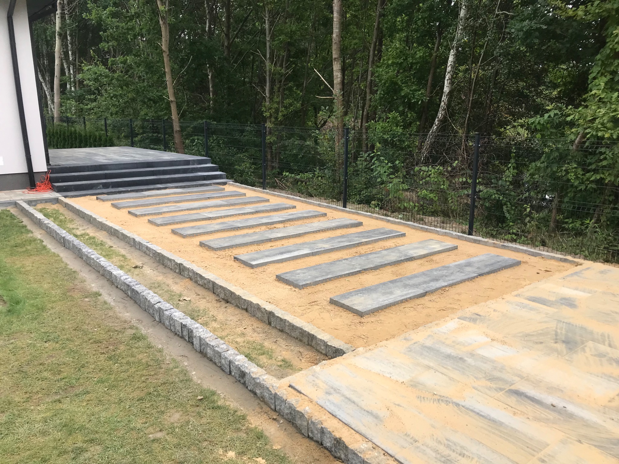 Przygotowanie ścieżki z prostokątnych płyt betonowych na piasku, obrzeża z kostki granitowej, widok na schody z kostki i zieleń ogrodu.