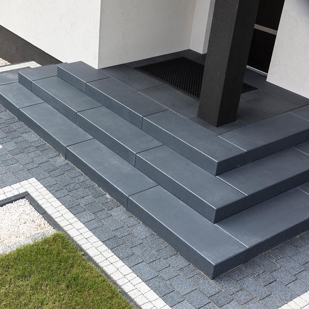 Nowoczesne schody wejściowe z szarej kostki brukowej, zintegrowane z chodnikiem z kostki o mniejszym formacie, obrzeże z białego kamienia, trawnik w lewym dolnym rogu, czarny słupek podtrzymujący...