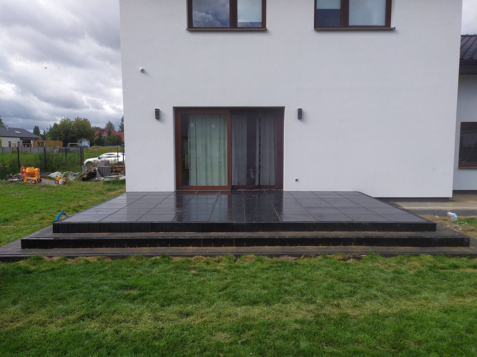 Wykończony taras z ciemnej kostki brukowej przy białym domu z przesuwnymi drzwiami tarasowymi, widoczne dwa stopnie i trawnik.