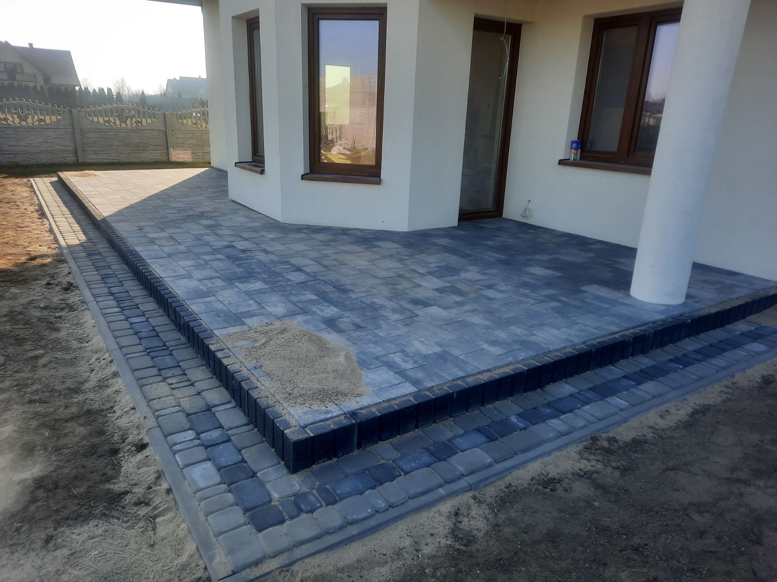 Nowo ułożona, szara kostka brukowa tworząca taras przy domu z widocznymi trzema schodkami i piaskiem na powierzchni, gotowa do fugowania.