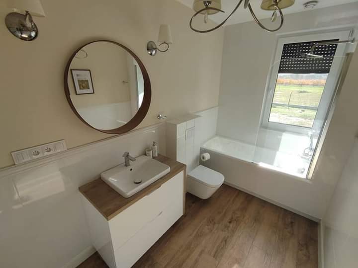 Jasna łazienka z drewnianą podłogą, okrągłym lustrem, umywalką na szafce z blatem imitującym drewno, toaletą w zabudowie i wanną pod oknem dachowym z roletą.