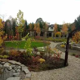 Jesienny krajobraz ogrodu z oczkiem wodnym, ścieżką z kamienia i drzewami o barwnych liściach, widok z poziomu gruntu.