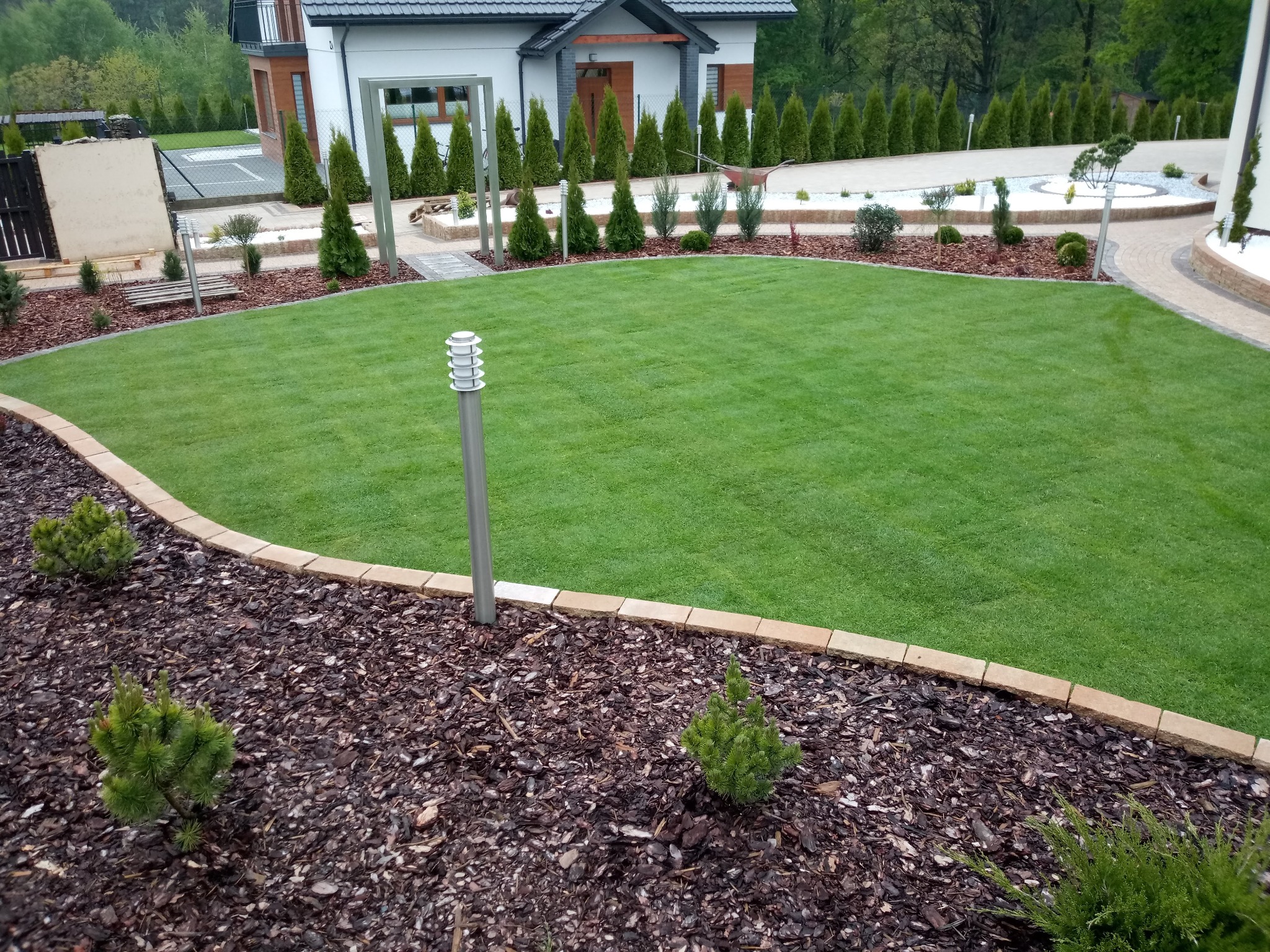Perfekcyjnie utrzymany trawnik z rolki, obrzeża wykończone kostką, rabaty wysypane korą z młodymi iglakami, nowoczesne oświetlenie ogrodowe i elegancki dom w tle.