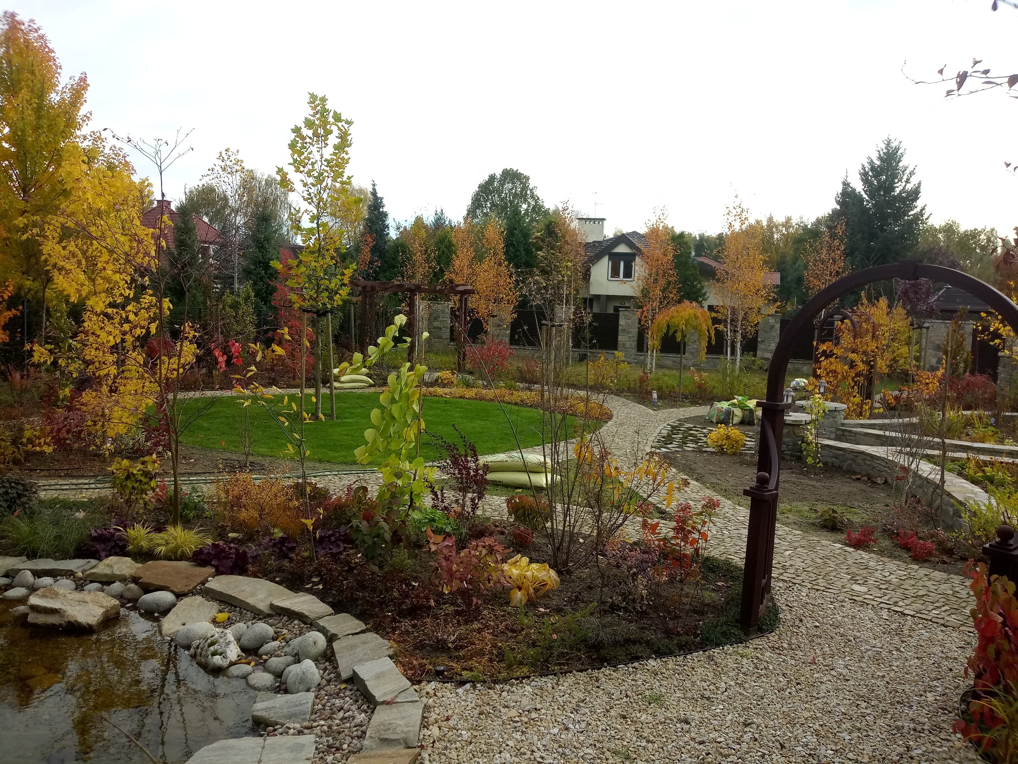 Jesienny krajobraz ogrodu z oczkiem wodnym, ścieżką z kamienia i drzewami o barwnych liściach, widok z poziomu gruntu.