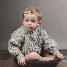 Kamila Jankowska Fotografia - Mała dziewczynka w grubym swetrze i opasce z kokardką siedzi w drewnianej misce na jutowym materiale, portret w neutralnej tonacji.